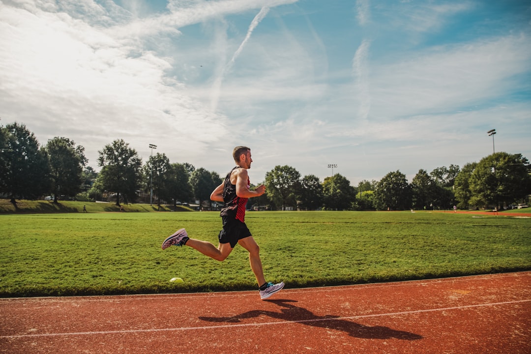 A man running on a red track beside a green field under blue sky